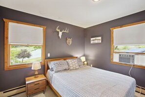 A bedroom with a bed, two windows, a nightstand, a lamp, a wall-mounted heater, and a wall decoration.