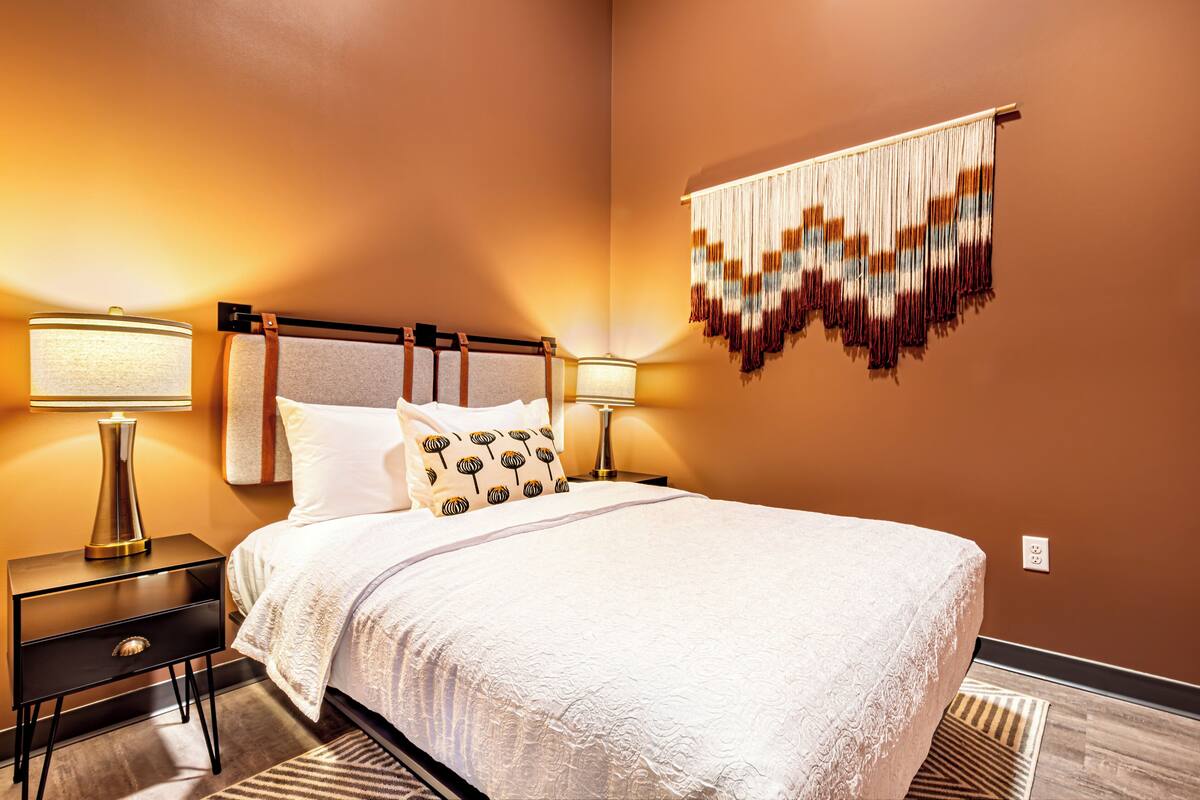 A neatly made bed with a white bedspread and patterned pillows, flanked by two bedside tables with lamps, in a room with a warm wall color and a decorative hanging piece.