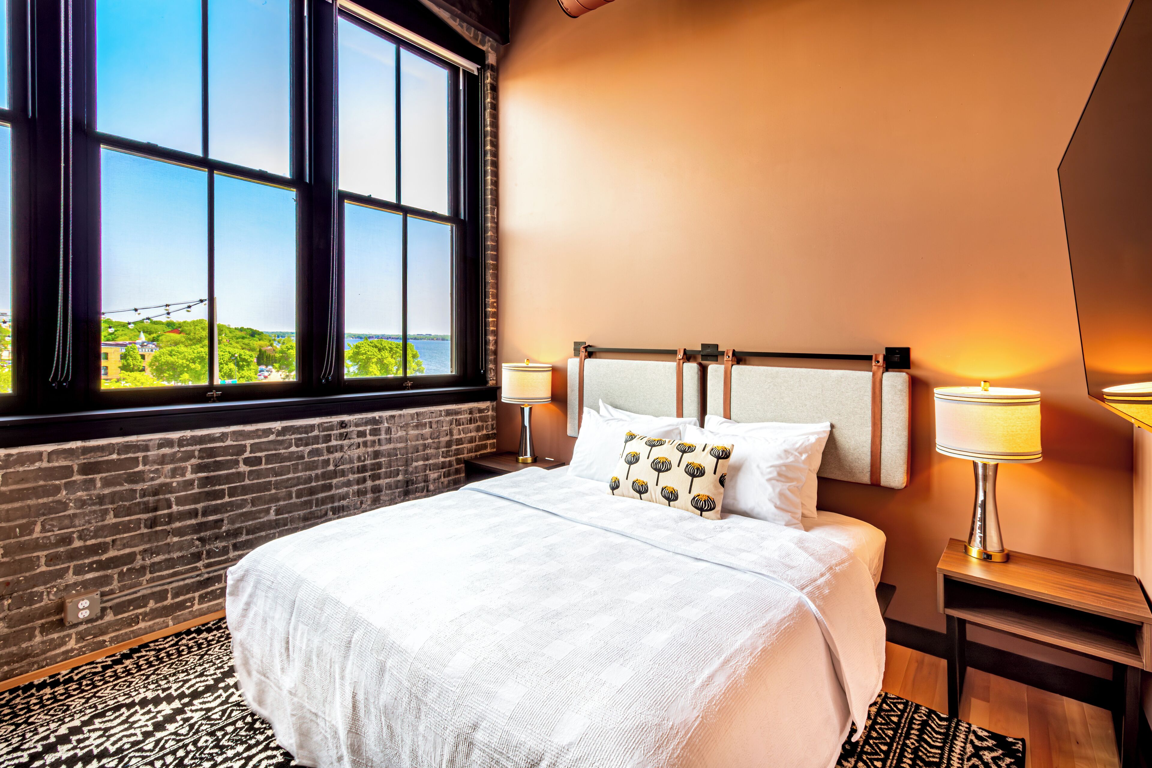 Habitación de hotel con un ventanal grande, una cama con ropa de cama blanca, dos lámparas de noche y una pared de ladrillo.