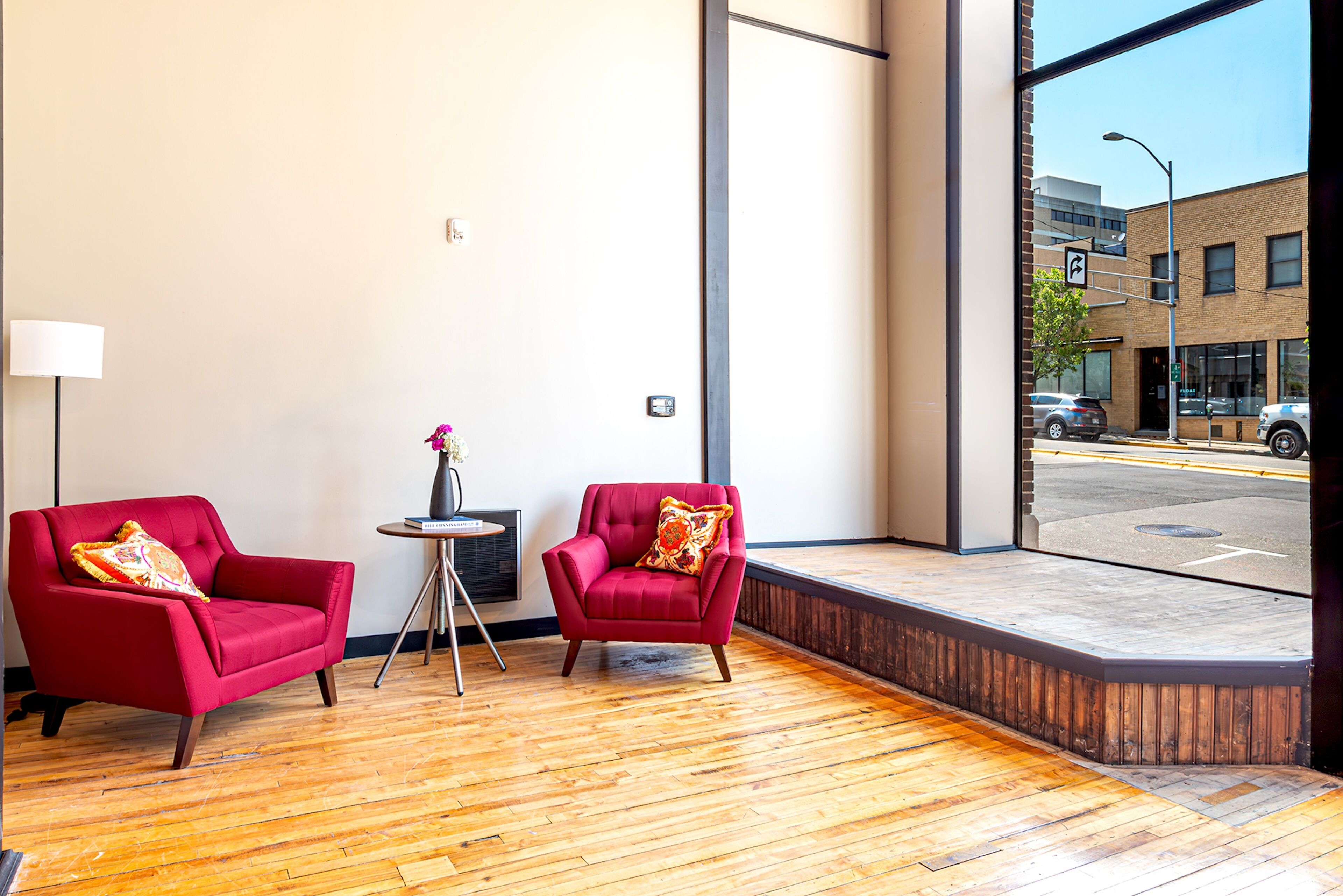 A room with two red armchairs, a small round table, and a large window offering a view of the street outside.