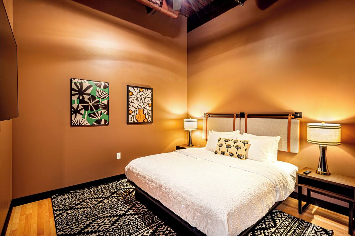 A hotel room with a large bed, two wall art pieces, a nightstand with a lamp, and a patterned rug.