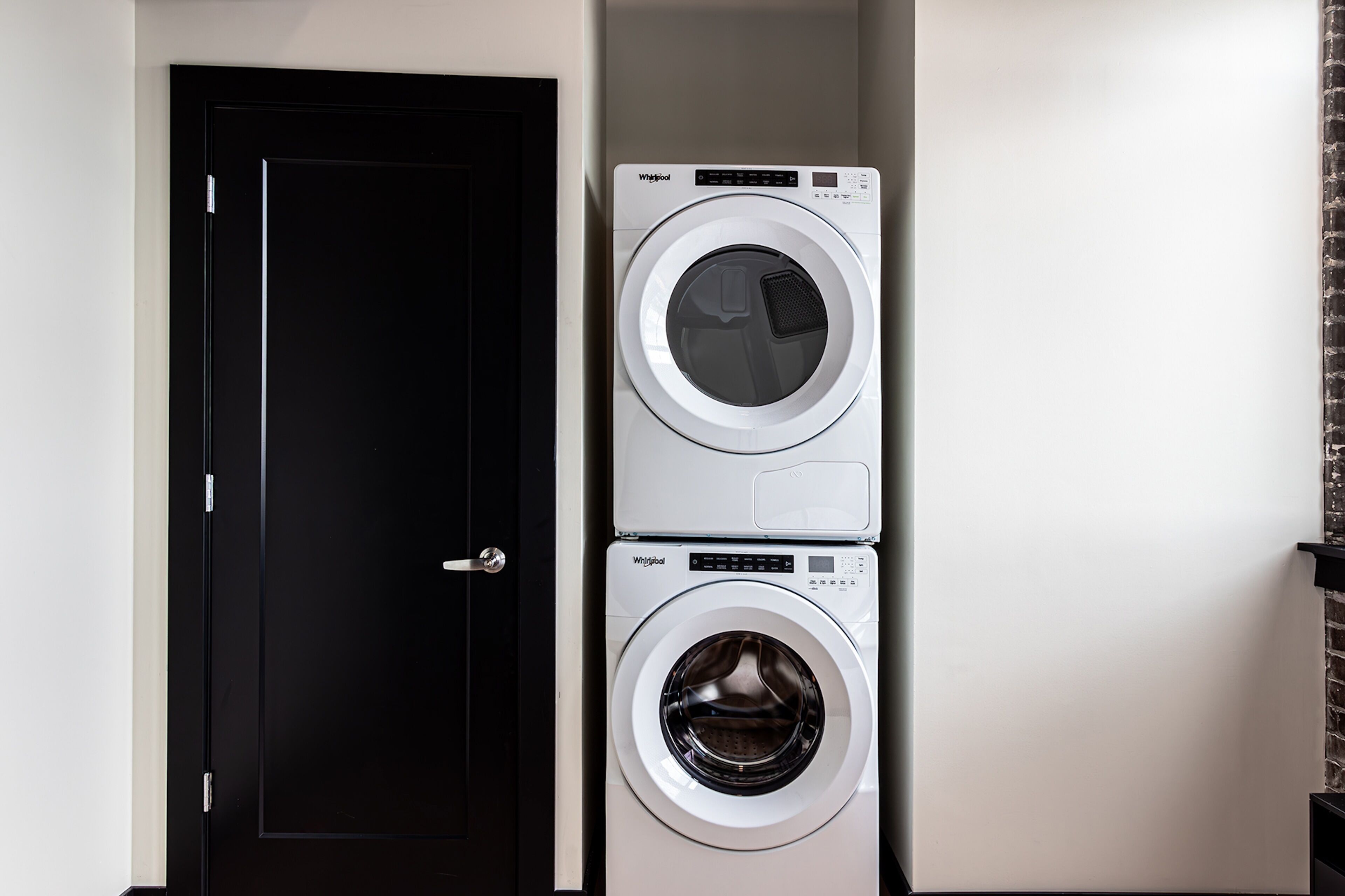 A stacked washer and dryer unit in a laundry room with a black door and a brick wall.