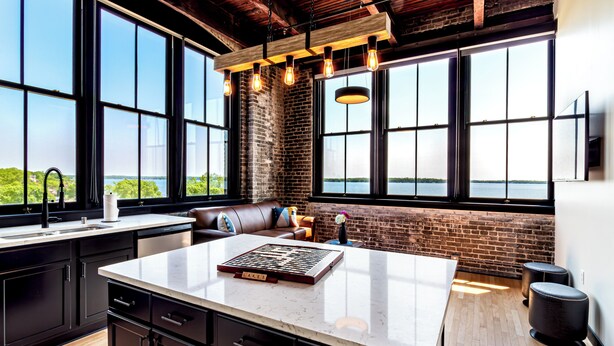 A modern kitchen with a large island, black cabinetry, and a view of a body of water through large windows. - The Saddlery Madison Downtown (Madison)