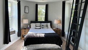 Une chambre à coucher avec un lit, deux tables de chevet, un ventilateur de plafond, une fenêtre et une échelle noire.