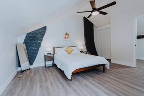 Ein Schlafzimmer mit schrägverdeckter Decke, einem Bett mit weißer Bettwäsche, zwei Nachttischen, einem Wandbild und einer Tür rechts.