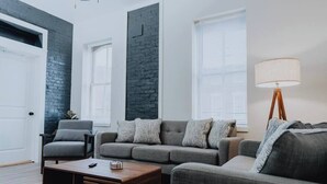 Ein modernes Wohnzimmer mit grauem Sofa, Couchtisch aus Holz, einer Stehlampe und einer Wand aus Ziegelsteinen.