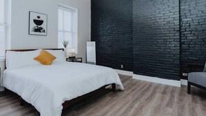 Ein Schlafzimmer mit einem Bett, einem Nachttisch, einer Lampe, einem Stuhl, einer schwarzen Ziegelwand und einem Fenster.