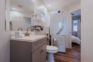 A modern bathroom with a white sink, mirror, and toilet. There is a small shelf with toiletries and a towel rack.
