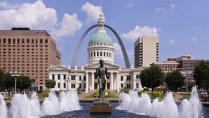 Ein Hotel mit einem großen Brunnen, einer Statue und einem markanten Gebäude mit Kuppel und Bögen im Hintergrund.