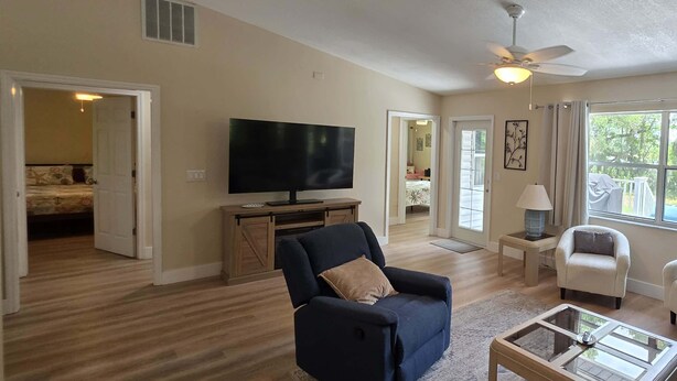 A spacious living room with a large flat-screen TV, a dark blue sofa, a glass coffee table, and a ceiling fan.