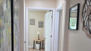 A hallway with a wooden floor, white walls, and a door leading to another room. There is a small table with a lamp and a framed picture on the wall.