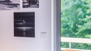 A room with a large window overlooking greenery, a wall-mounted light switch, and a framed photograph of a lighthouse.