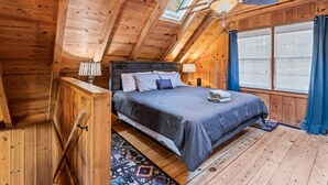 Un dormitorio de cabaña de madera con cama, ventilador de techo y una ventana grande con cortinas.