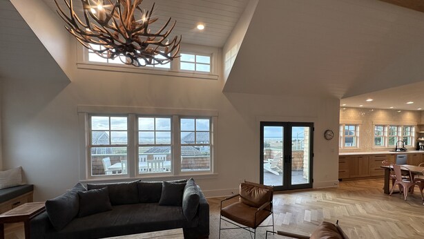A spacious living room with a large sectional sofa, a coffee table, and a chandelier. There are large windows offering views of the ocean and a patio area with outdoor seating. - The Ridge - Gearhart, OR (Gearhart)