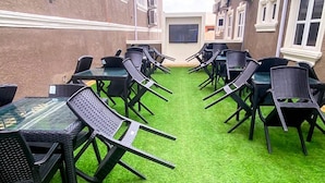 Un espace extérieur aménagé avec plusieurs tables et chaises, un téléviseur à écran plat fixé au mur et un sol recouvert d’un gazon synthétique vert.