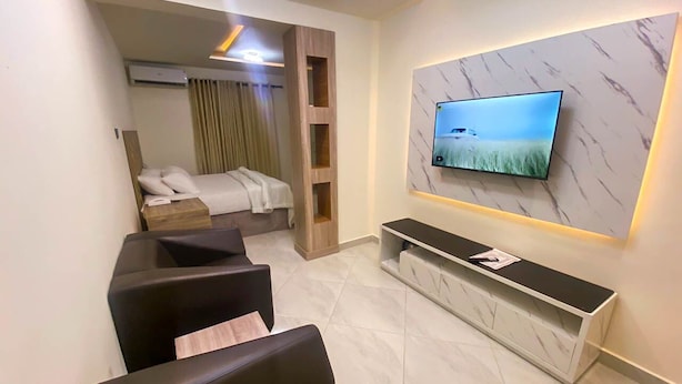 Une chambre d’hôtel moderne avec un téléviseur fixé sur un mur de marbre, un lit avec une tête de lit et un coin salon agrémenté d’une console.
