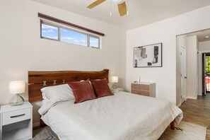 Ein Schlafzimmer mit einem Holzkopfende, einem großen Bett mit weißen und roten Kissen, einem Nachttisch mit Lampe sowie einem zusätzlichen Beistelltisch und einem Fenster mit Jalousien.