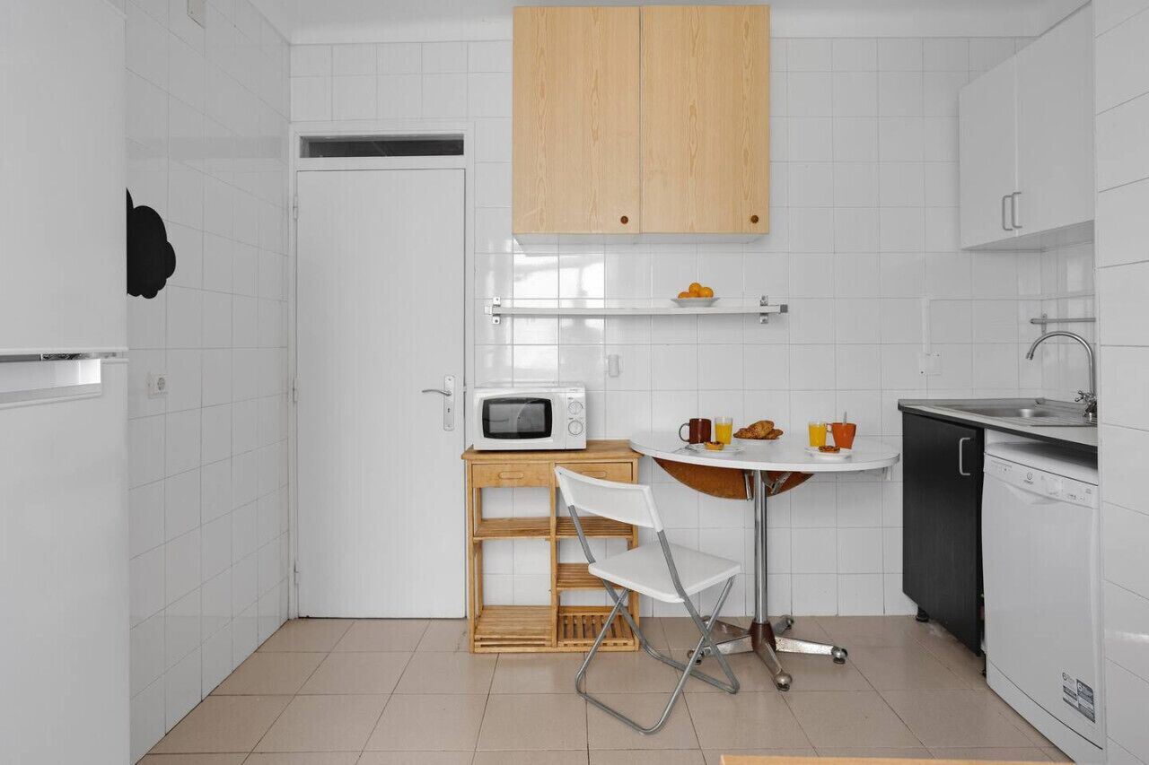 Una cocina compacta con armarios blancos, un microondas, una mesita de madera con una silla y un fregadero.