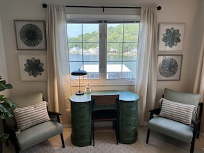 A room with a desk, two chairs, a lamp, and a view of a body of water through a window.