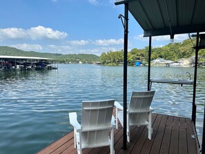 Eine Holzsteg mit weißen Stühlen und einem überdachten Bereich mit Blick auf einen ruhigen See, im Hintergrund Boote und Häuser.