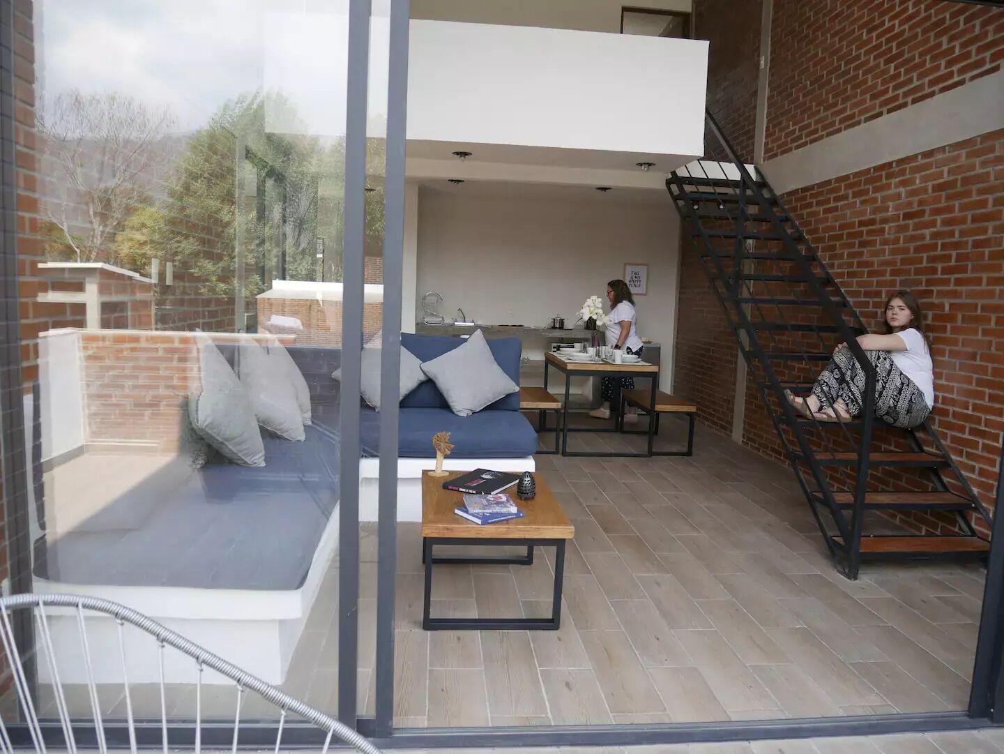 A modern loft with a glass wall, a blue sofa, a wooden table, and a staircase.