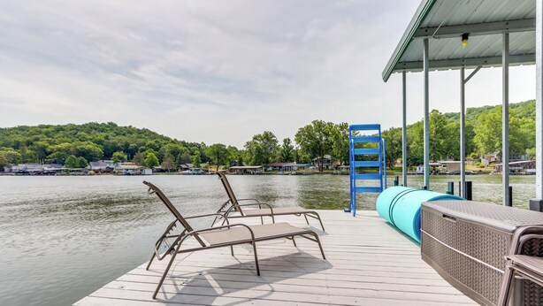 A wooden dock with lounge chairs and a covered seating area overlooking a lake with houses in the background. - Fire Pit & Dock: Lake of the Ozarks Retreat! (Roach)