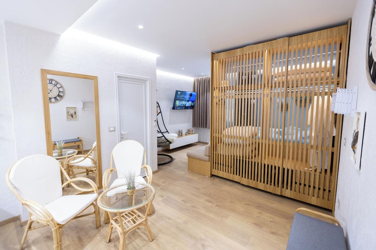 A modern living room with a glass table, two white chairs, a wooden partition, a TV mounted on the wall, and a wooden floor.