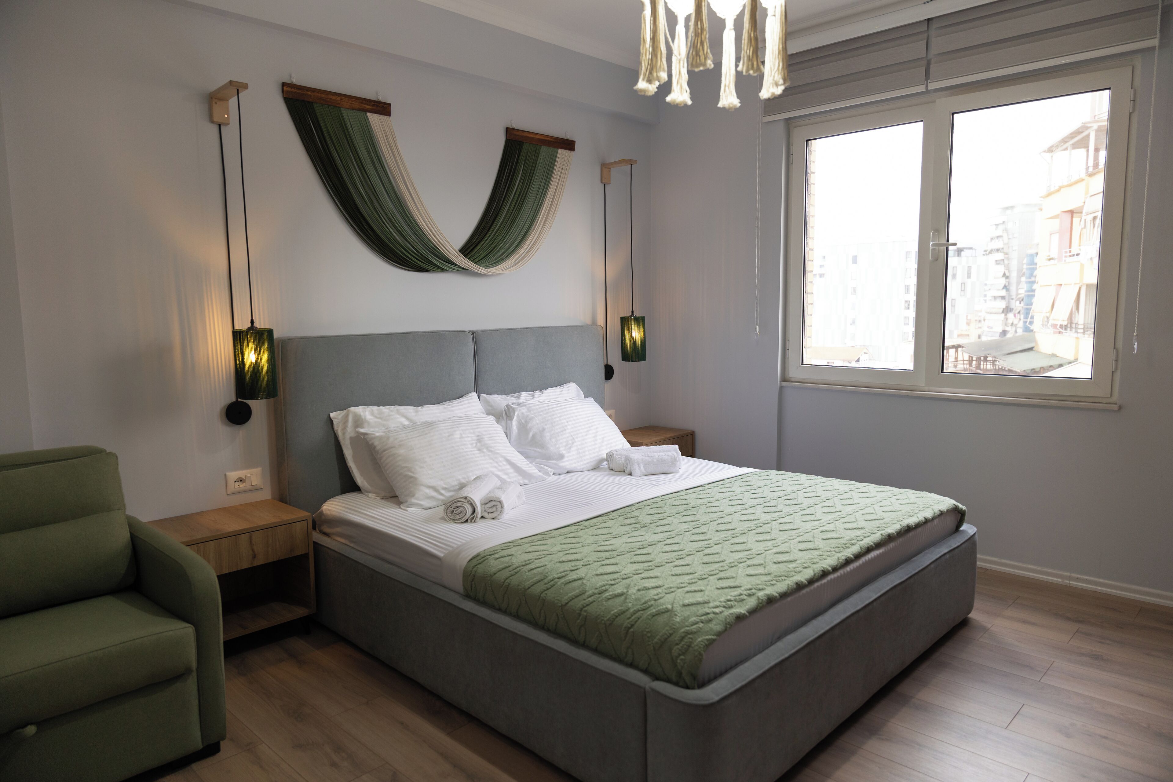 A modern bedroom with a large bed, two bedside tables, a green armchair, and a window with a city view.