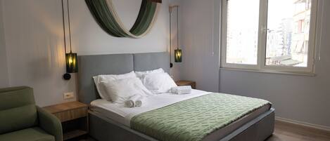 A modern bedroom with a large bed, two bedside tables, a green armchair, and a window with a city view.