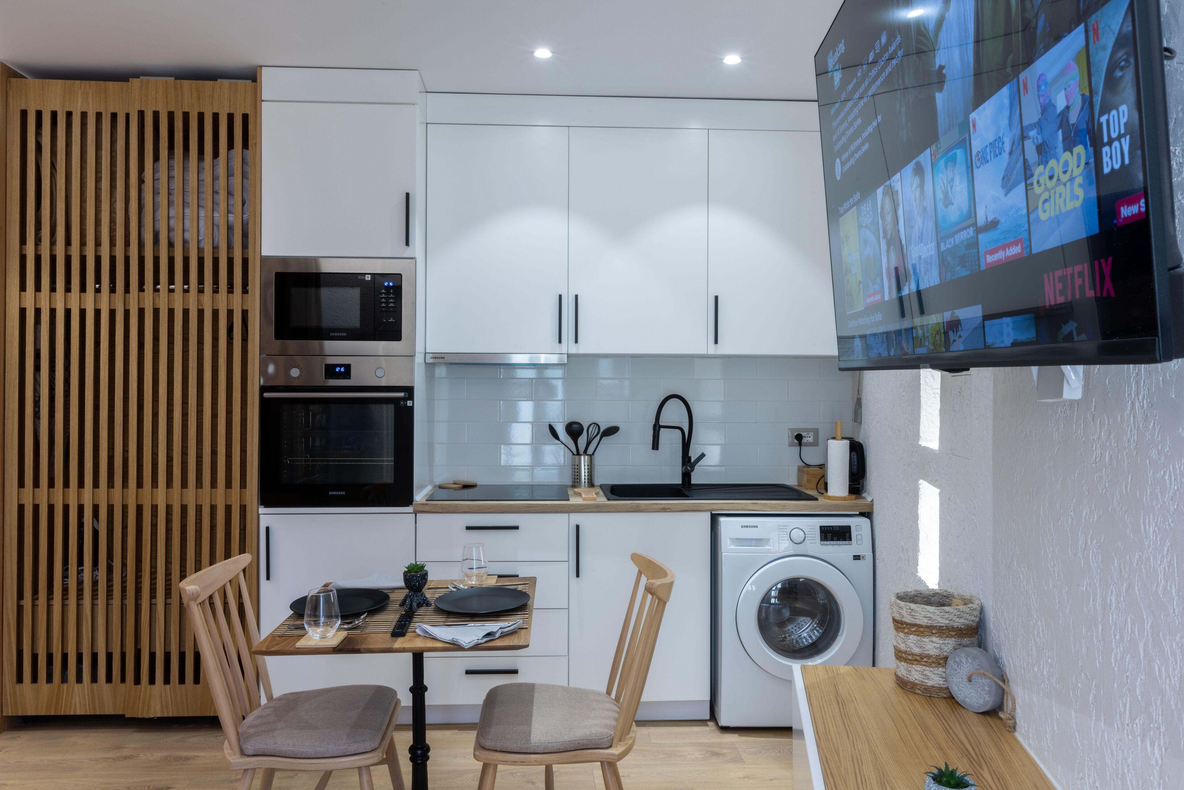 A modern kitchen with white cabinets, a microwave, an oven, a sink, and a washing machine. There is a dining table with chairs and a TV mounted on the wall displaying a streaming service interface.