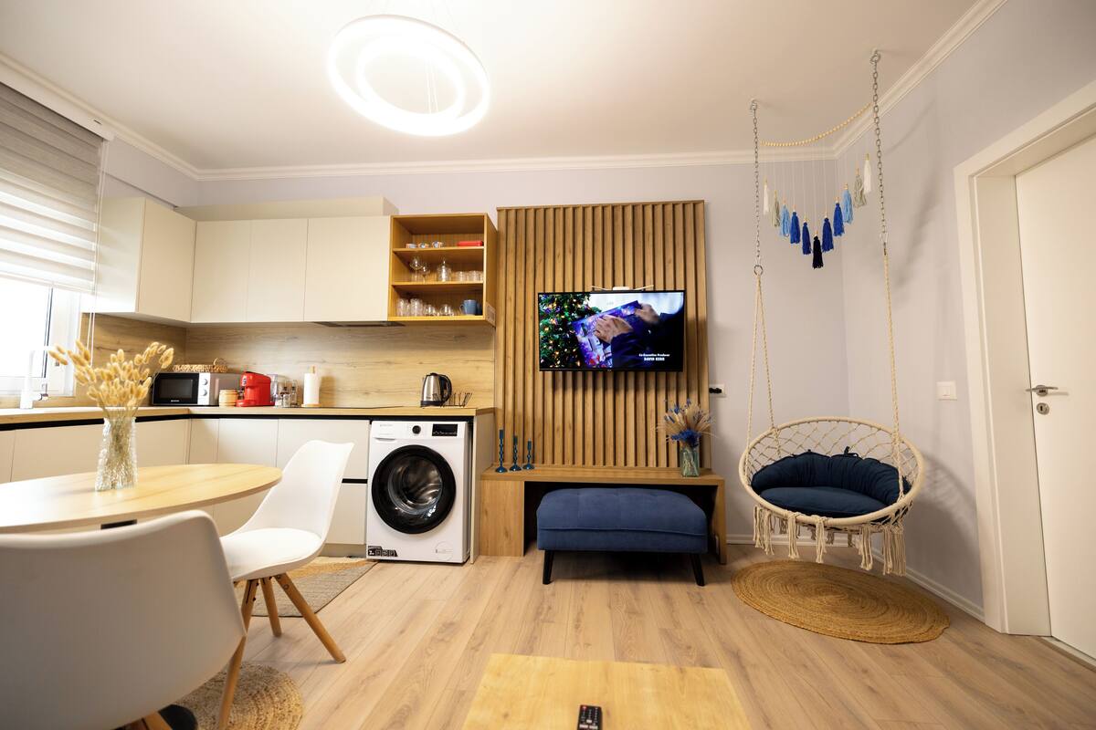A modern living room with a washing machine, a television, a dining area, and a hanging chair.