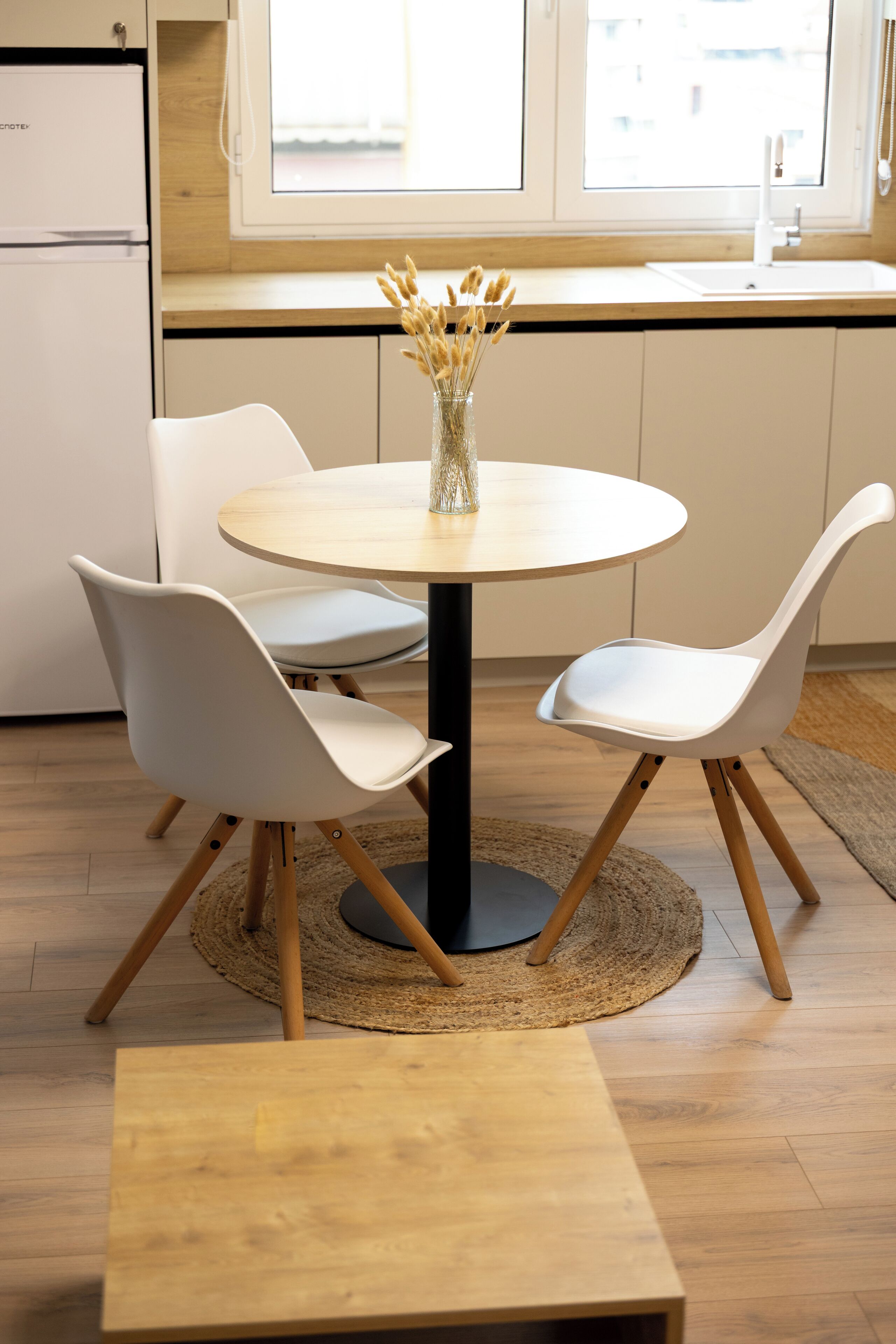 A modern kitchen with a round dining table, white chairs, and a vase of dried flowers.
