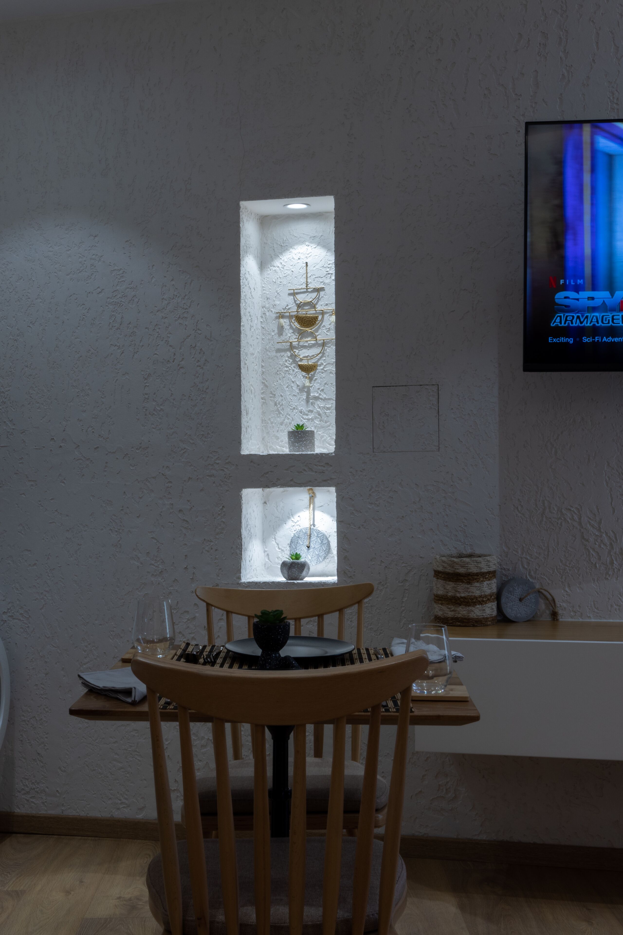 Un coin repas avec une table et des chaises en bois, une petite plante en pot et un téléviseur fixé au mur diffusant un film.
