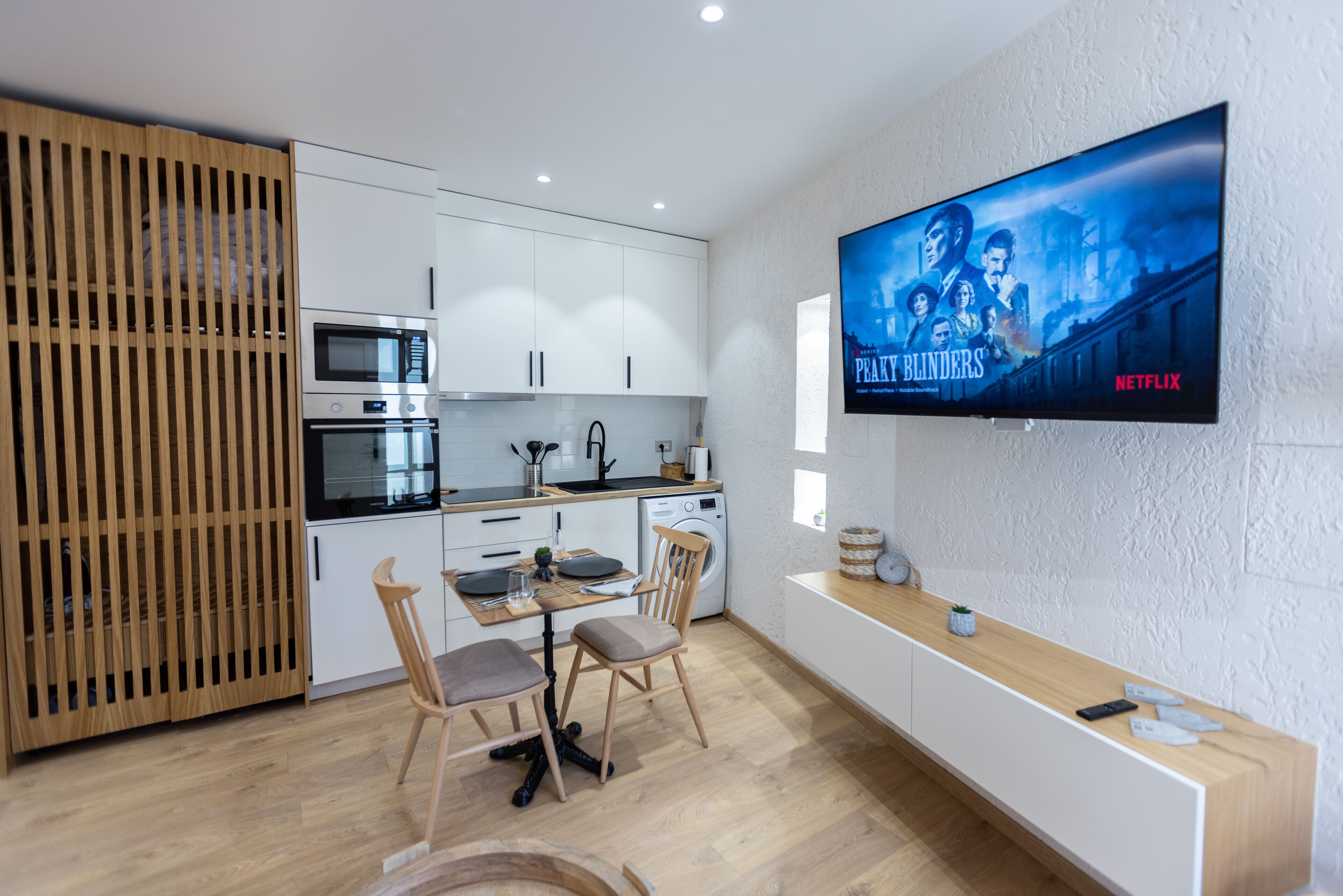 A modern kitchen with white cabinets, a wooden dining table, and a TV displaying 'Peaky Blinders' on Netflix.