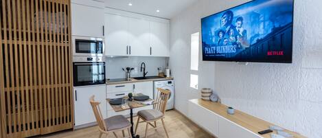 Cocina moderna con armarios blancos, una mesa de comedor de madera y un televisor que muestra 'Peaky Blinders' en Netflix.