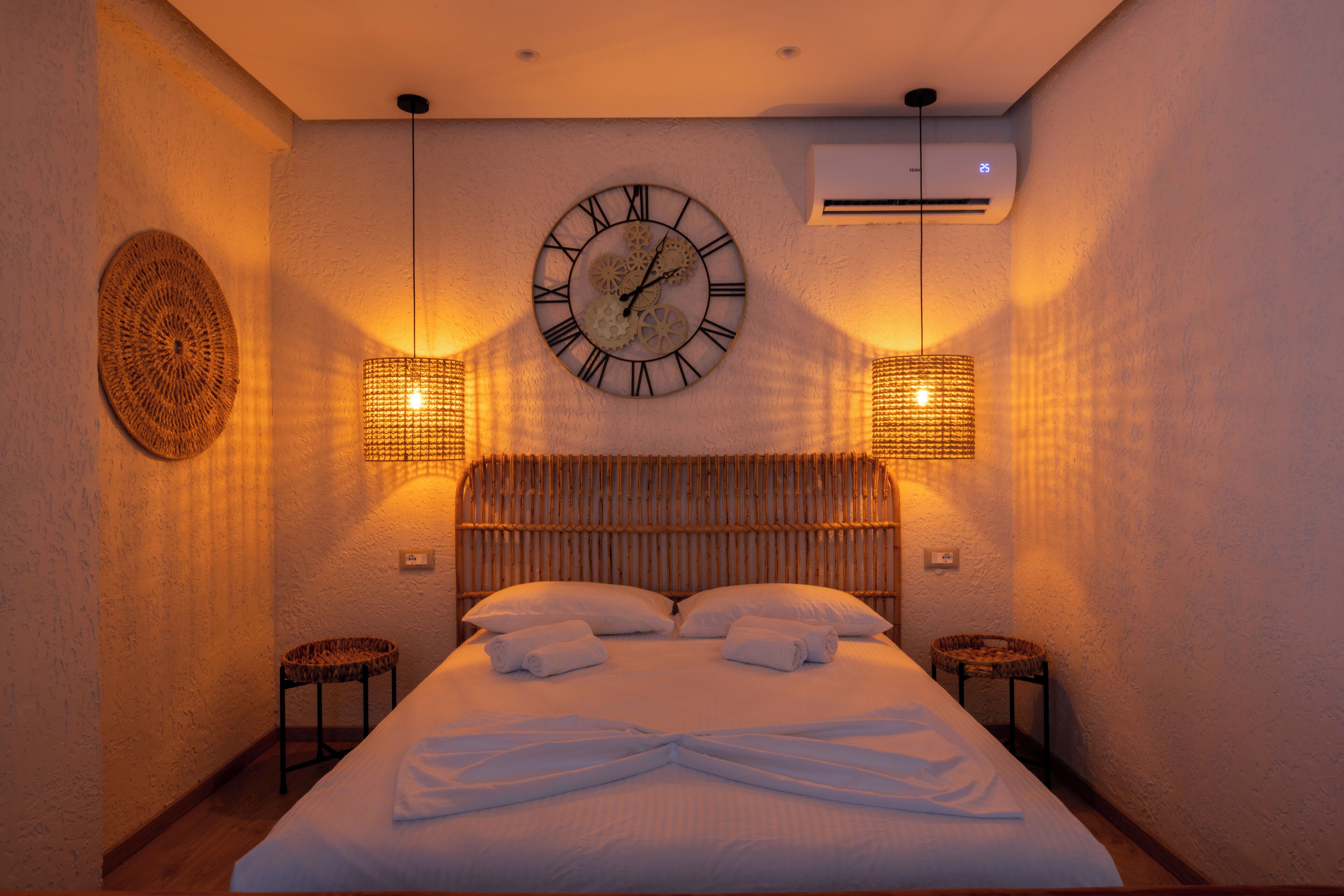 A neatly made bed with white linens, a round wall clock, and two wall-mounted lamps.