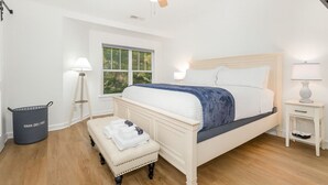 Un dormitorio con cama, un banco, ventilador de techo, ventana y un cubo de basura.