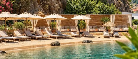 A pool area with lounge chairs and umbrellas, surrounded by greenery and a rocky cliff.