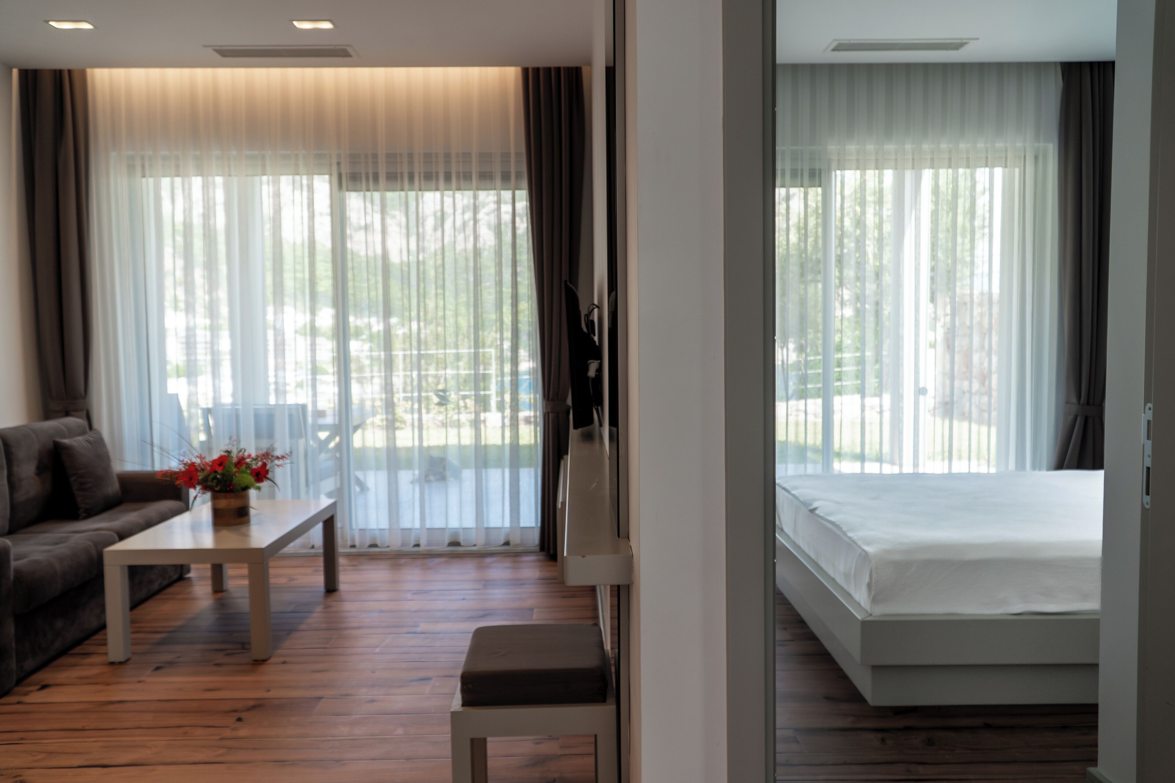 Habitación de hotel moderna con suelo de madera, un sofá, una mesa de centro con flores, una cama y ventanas grandes con cortinas traslúcidas.