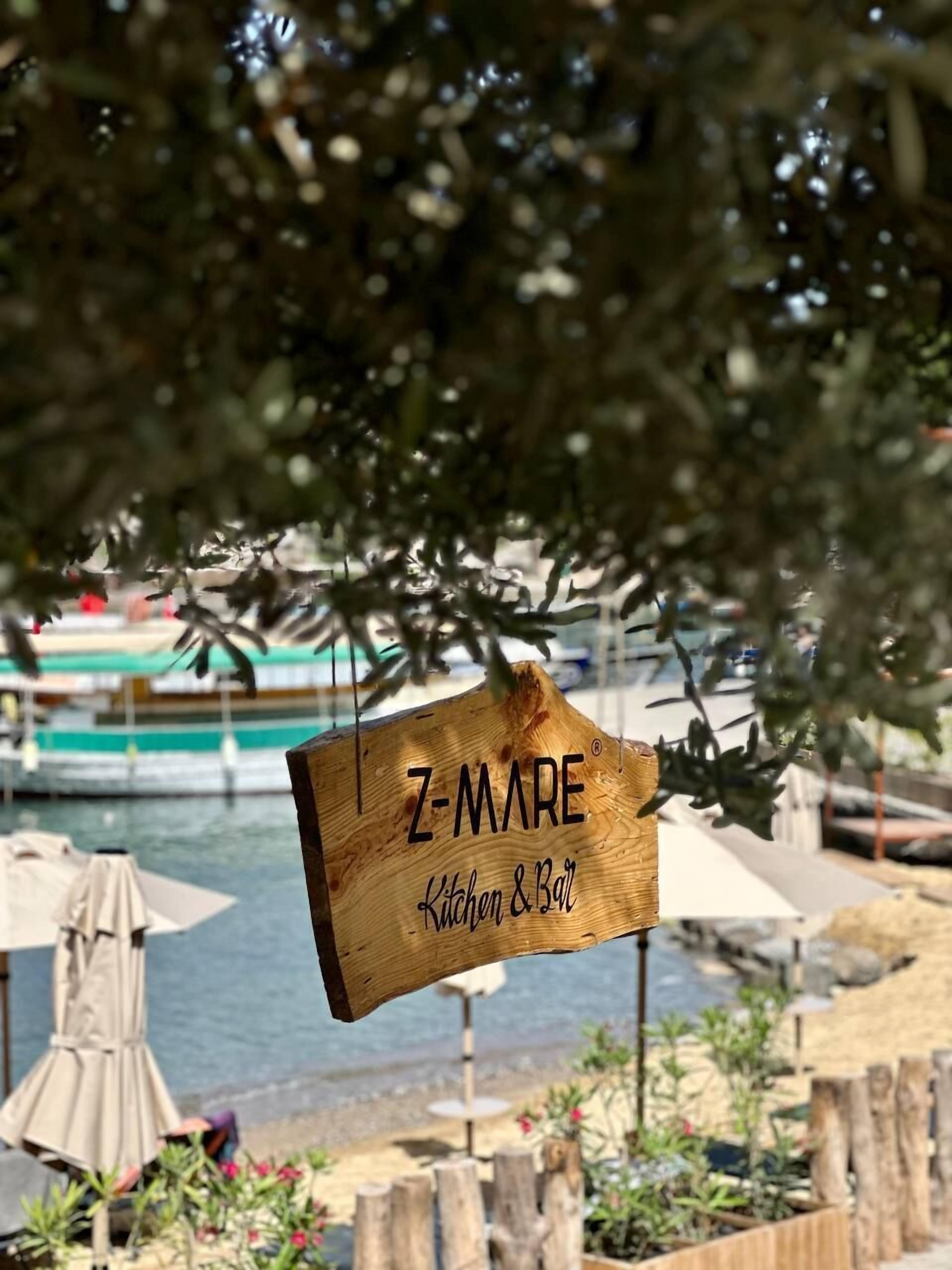 A wooden sign with "Z-MARE Kitchen & Bar" written on it, set against a backdrop of a beach with umbrellas and a body of water.