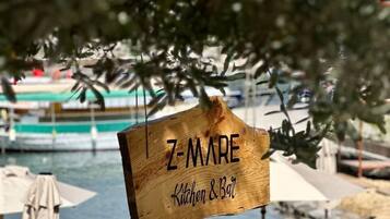 A wooden sign with "Z-MARE Kitchen & Bar" written on it, set against a backdrop of a beach with umbrellas and a body of water.