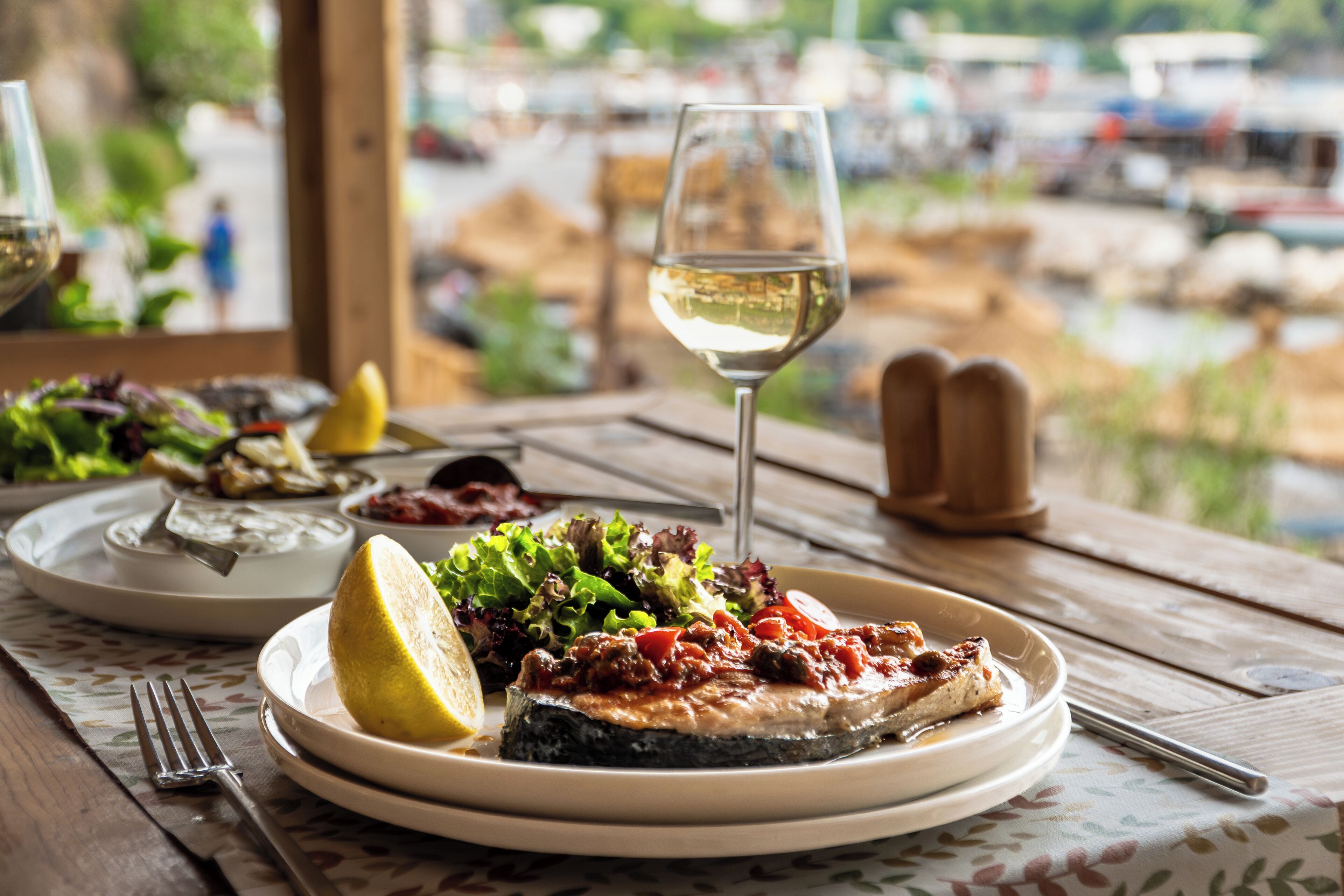 A dining table with plates of food, a glass of white wine, and a view of the outdoors.