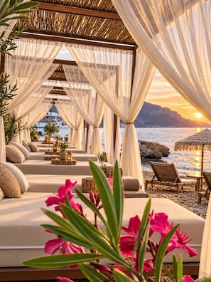 A beachside lounge area with cushioned seating, a view of the sea, and a sunset in the background.