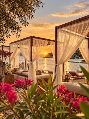 A beachside lounge area with a view of the sunset over the water, featuring a canopy bed and lounge chairs.