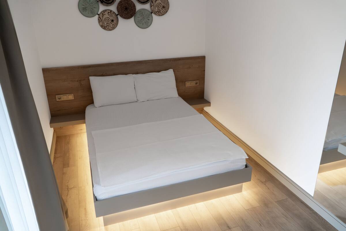 A modern bedroom with a wooden headboard, a neatly made bed with white linens, and a wall-mounted decorative plate above the bed.