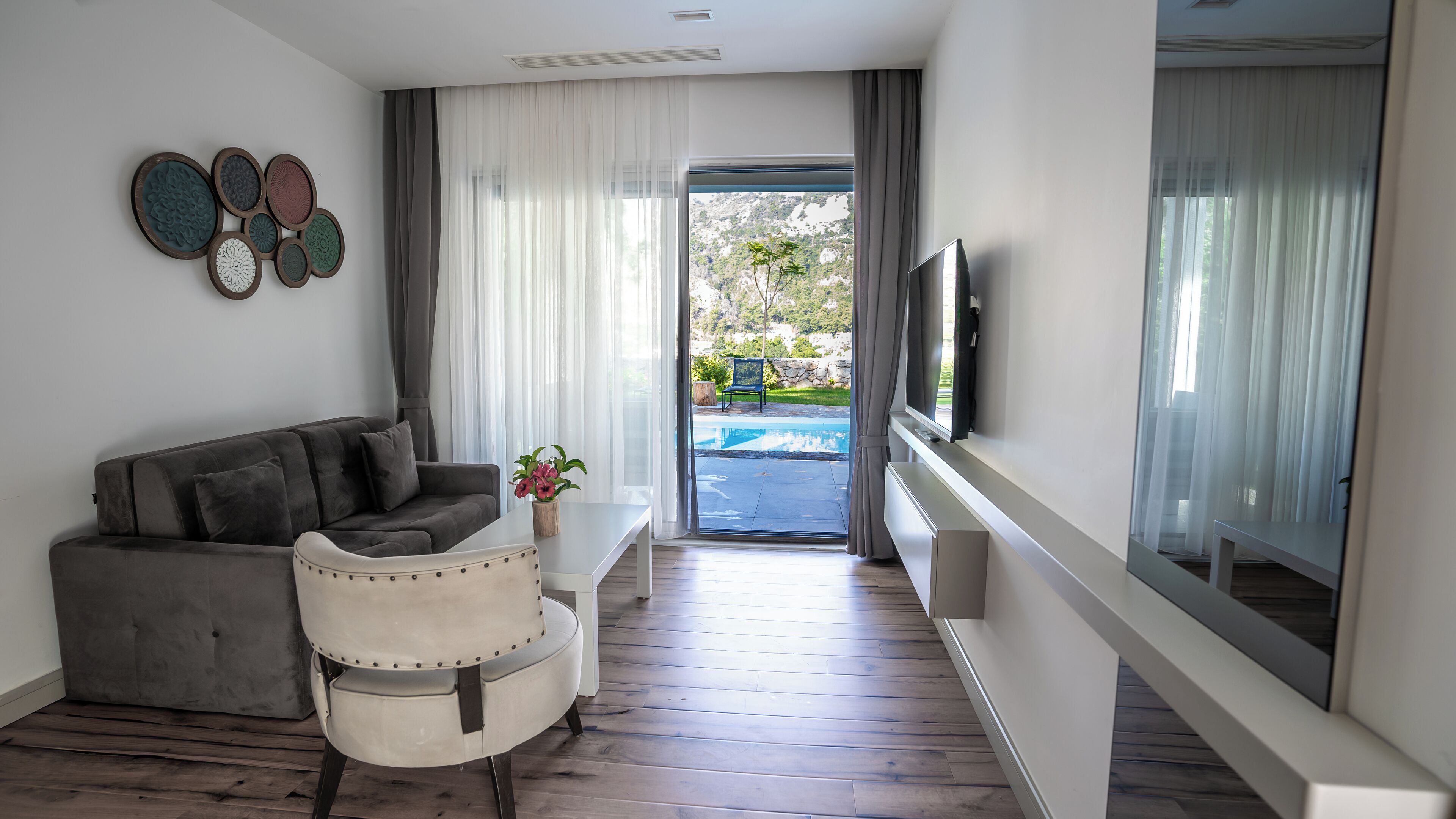 Una sala de estar moderna con un sofá gris, un sillón blanco, una mesita con una planta y un televisor montado en la pared.