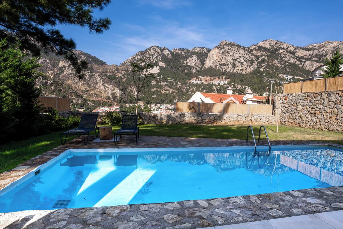A swimming pool with stone edging, two lounge chairs, and a stone wall.