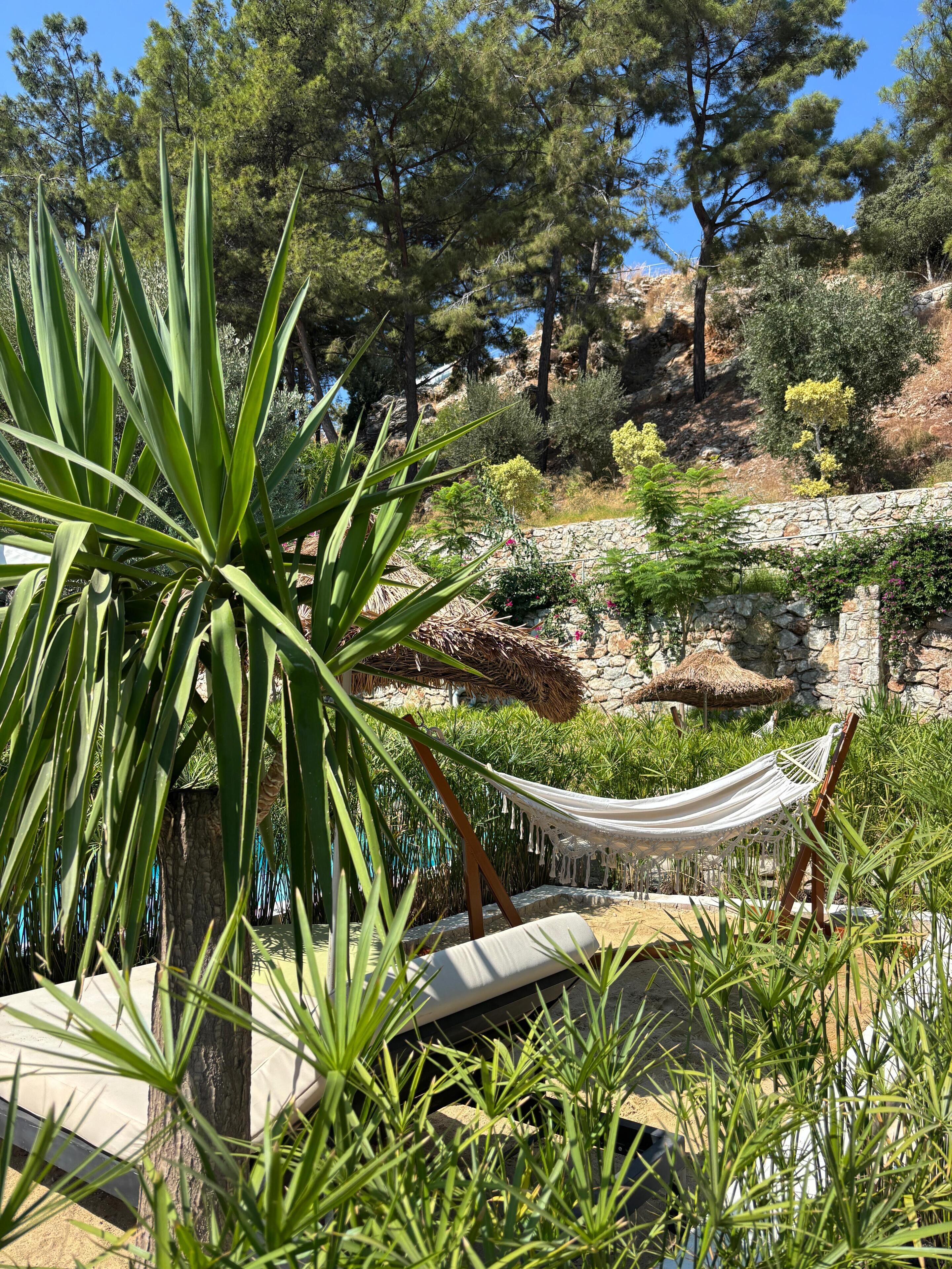 A hammock in a lush garden with a stone wall and a rocky hillside in the background.