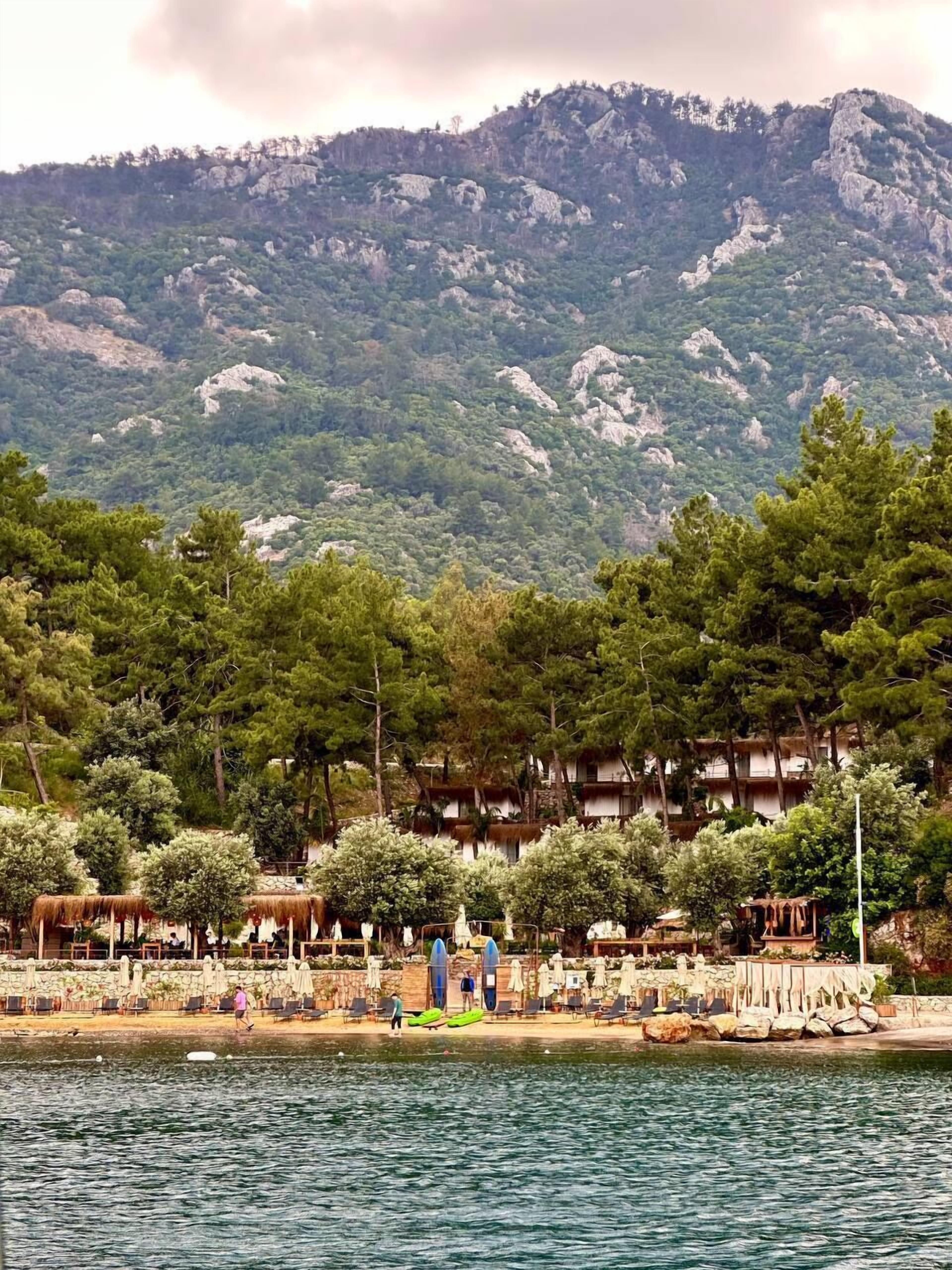 A coastal hotel with outdoor seating and umbrellas, overlooking a body of water and a mountainous backdrop.