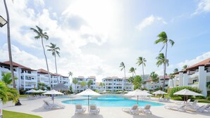 A resort pool area with lounge chairs and umbrellas, surrounded by palm trees and hotel buildings. - Just  few steps from Beach, Enjoy a Sunrise in this Wonderful place (Punta Cana)